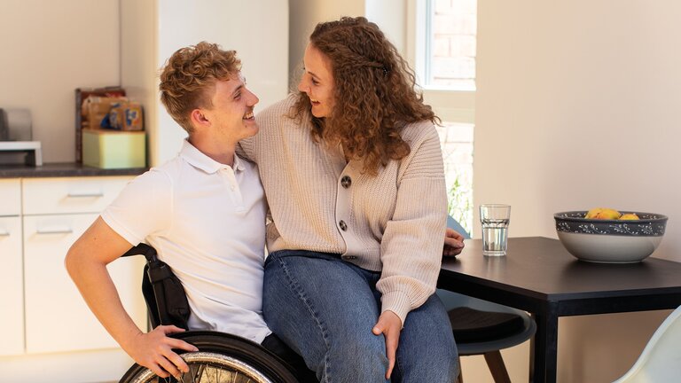 Eine junge Frau sitzt auf dem Schoß eines jungen Mannes, der im Rollstuhl sitzt an einem Tisch. Sie lächeln sich an.  | © ©adira.de, Fotograf: Daniel George