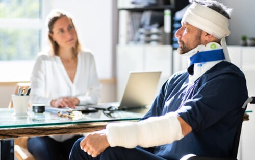 Ein Mann sitzt einer Ärztin im Sprechzimmer gegenüber. Der Mann sitzt im Rollstuhl und trägt eine Nackenstütze, Kopfverband und einen Gips am linken Arm. | © Unsplash