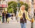 Eine junge Frau mit einem Blindenstock überquert eine belebte Straßenecke. Sie trägt eine gelbe Jacke, ein dunkelbraunes Oberteil und blaue Jeans. Ihr Gesichtsausdruck wirkt konzentriert. Im Hintergrund sind einige Passanten, darunter ein Mann mit einer grünen Tasche und ein weiterer in dunkler Kleidung. Es gibt auch Straßenschilder und Graffiti. | © Gesellschaftsbilder / Andi Weiland