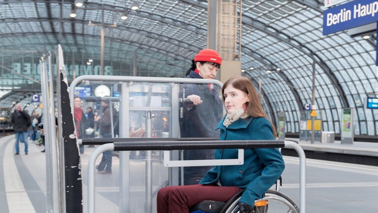 Im Vordergrund ist eine Frau im Rollstuhl zu sehen, die von einer Frau mit roter Mütze auf eine Hebebühne begleitet wird. Im Hintergrund ist ein blaues Schild mit der Aufschrift "Berlin Hauptbahnhof" zu sehen. Die Personen befinden sich auf einem Bahnsteig. | © Wolfgang Bellwinkel | DGUV