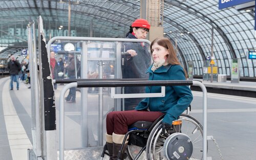 Eine Frau im Rollstuhl wird die von einer Bahnmitarbeiterin in einer roten Mütze auf einer Hebebühne begleitet. Sie befinden sich auf einem Bahnsteig im Berliner Hauptbahnhof. Im Hintergrund sind weitere Personen und die Struktur des Bahnhofs zu sehen. Ein Schild mit der Aufschrift "Berlin Hauptbahnhof" ist ebenfalls sichtbar. | © Gesellschaftsbilder - Wolfgang Bellwinkel | DGUV
